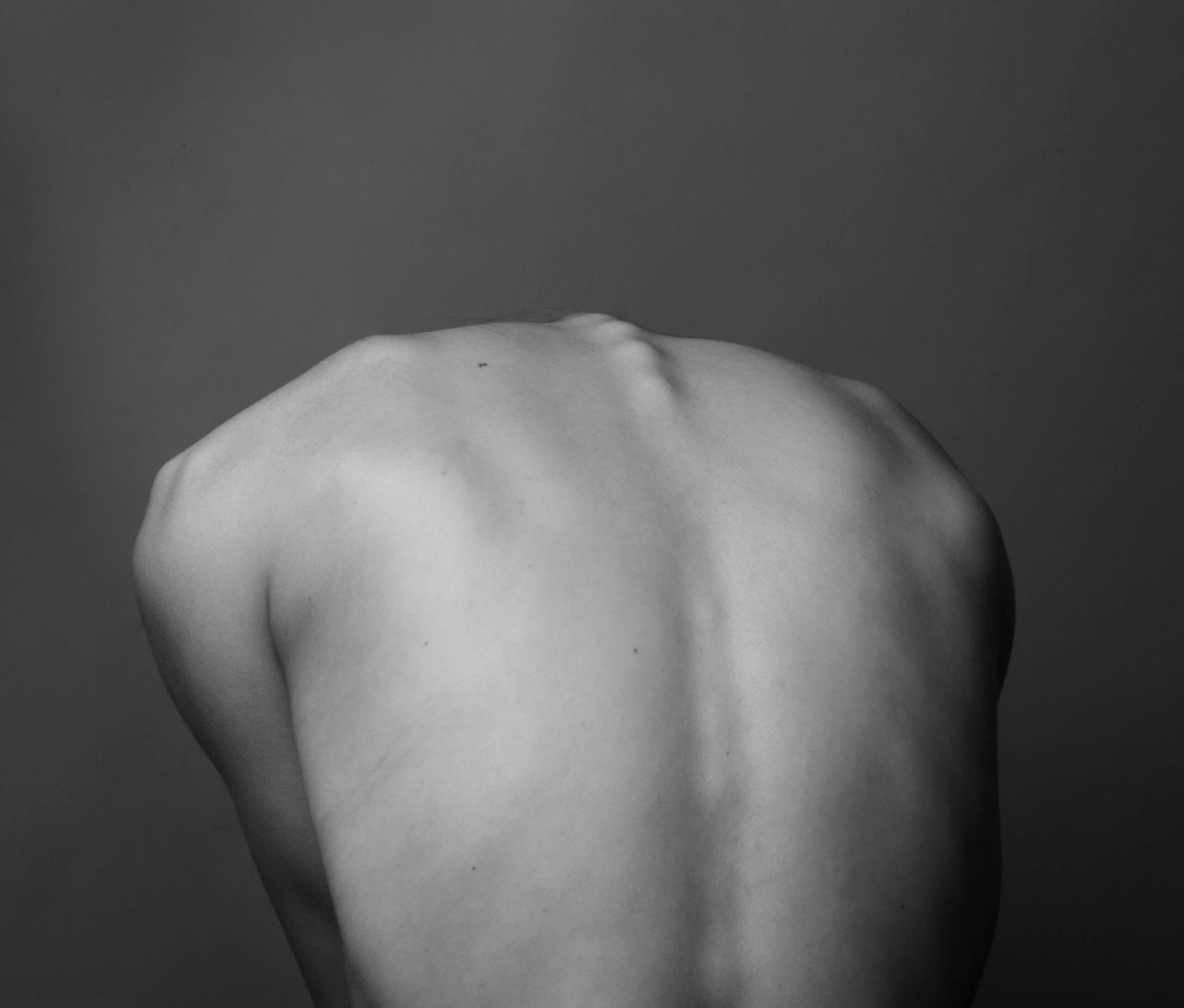 "Black and white photograph of a person's bare back showing the lower back and shoulder blade area against a dark background"